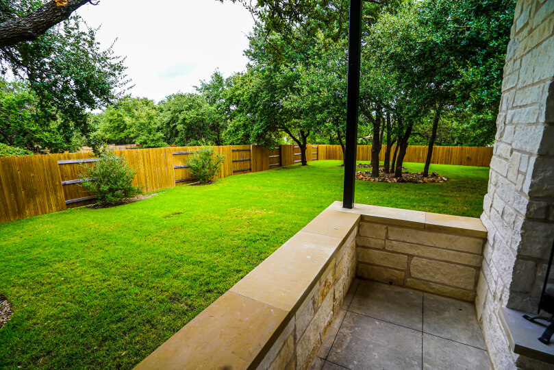 View looking out over a low stone wall and out toward lush green grass. A light wooden fence surrounds the perimeter, and there are small bushes and several trees.