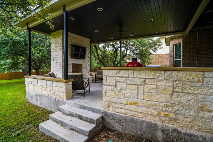 3 cement steps leading up to a covered light stone deck with a fireplace and a TV mounted above it.