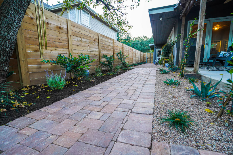 Light brick path with plants surrounded by gravel on one side, and larger plants in mulch along a light fence on the other side.