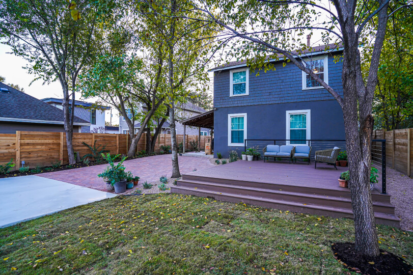 View of a backyard looking toward the back of a blue house. There's a dark wooden deck next to the house and a red brick patch next to the deck.