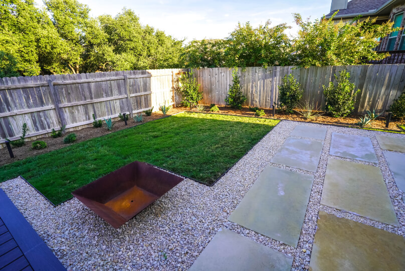 Raised bed with gravel and rectangular stone pavers next to a patch of lush green grass. Around the perimeter of the yard, there's a light wooden fence with small plants lining the inside.
