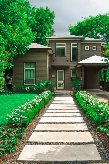 Front view of a walkway leading up to a front door. There are white stone steps leading up to a brown house. White flower shrubs line the pathway and green grass can be seen in the background.
