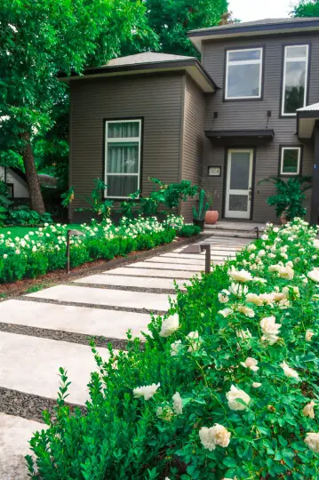 Front entrance to a house. There are white stone steps leading up to the front porch and there are flower plants on either side of the path.