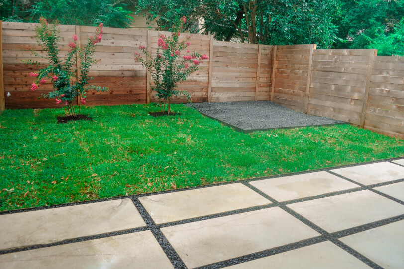 A backyard with white stone landscaping and grass. There is a fire pit and two pink flower bushes are in the background.