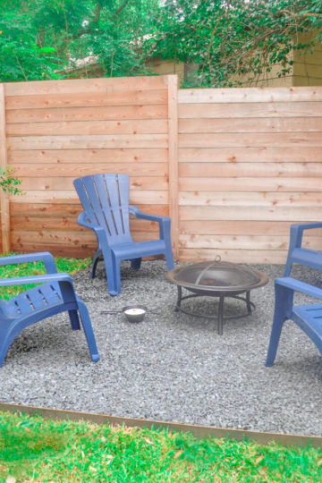 Backyard firepit with four plastic chairs. The fire pit is surround by grey gravel. A wooden fence can be seen in the background.