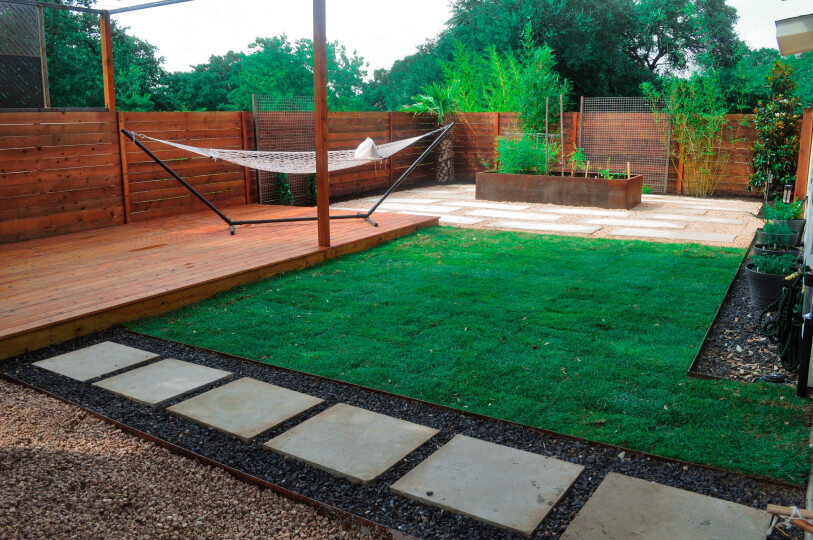 View of a landscaped yard. There is white stepping stones leading to a wooden patio with a hammock on it. You can see grass and gravel in the background.