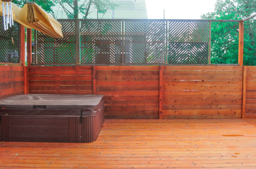Close up of a wooden patio in a backyard. On the patio there is a covered hot tub. Behind it is a wooden and metal fence.
