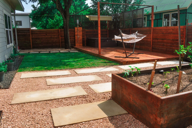 A landscaped yard with gravel and white stepping stones. There is a grass and wooden patio with a hammock on it. You can see a raised plant bed with small green plants.