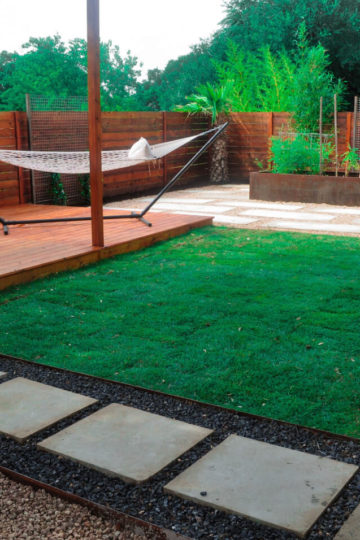 Landscaped backyard with a wooden patio with a hammock on it. There is white stepping stones in dirt. There is grass and gravel in the background. The backyard is surrounded by a wooden fence.