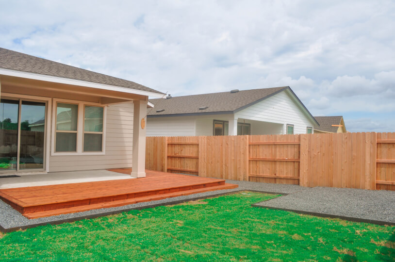 Wide view of a backyard where the back of the house is seen. The backyard features grass and grey gravel. The house next door can be seen behind the wooden fence.
