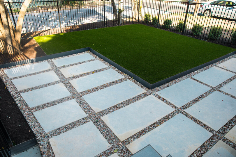 Small backyard with a patch of lush green grass next to an area with white rectangular pavers lined with gravel.