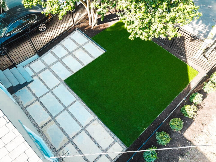 Aerial view of a small backyard with concrete stairs coming off a house leading to an area of the yard tiled with white rectangular pavers lined with gravel. Next to the paved area, there is a patch of lush green grass.