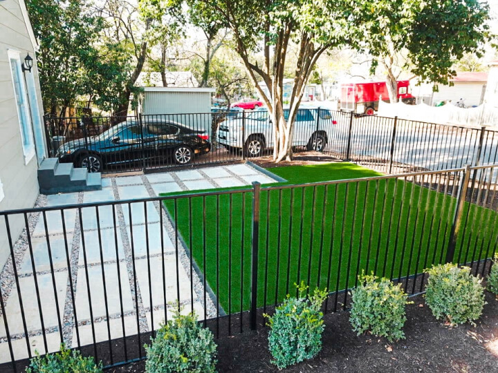 Small backyard with a black iron fence around the perimeter. There are small shrubs lining the outside of the fence. Inside the fence, a patch of lush green grass next to an area with white rectangular pavers lined with gravel.