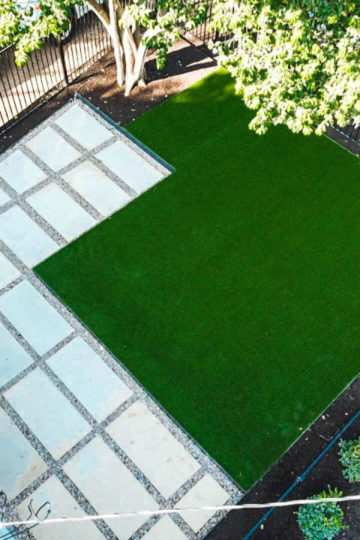 Aerial view of a small backyard with concrete stairs coming off a house leading to an area of the yard tiled with white rectangular pavers lined with gravel. Next to the paved area, there is a patch of lush green grass.