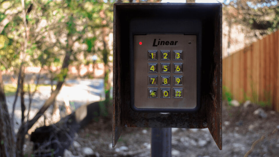Gate code keypad with metal cover. Trees can be seen in the background.