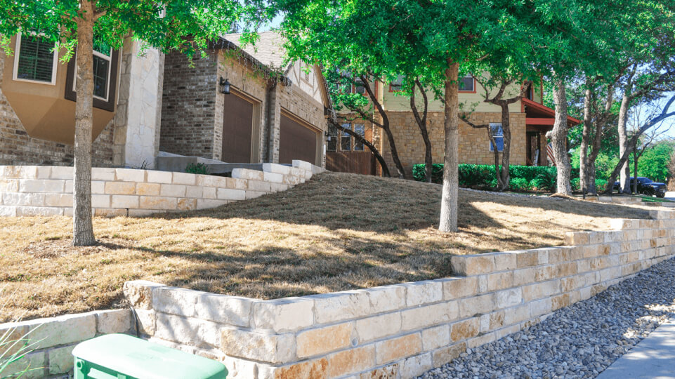 Side view of a front yard. Front yard is surround by short stone wall. Grass and trees are seen in the background.