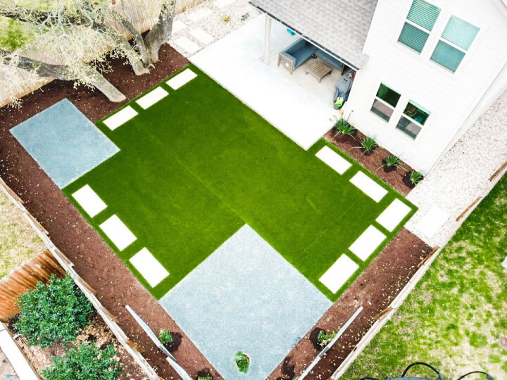 Top view of a landscaped backyard. There is a grass with white stepping stones and two grey pebble gardens. You can also see the top of the back of the house and the trees in the backyard.