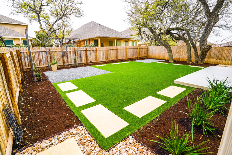 View of a landscaped backyard. There is a rectangular patch of grass with white stepping stones. There is a wooden fence surrounding the yard. You can see trees and a house in the backyard.