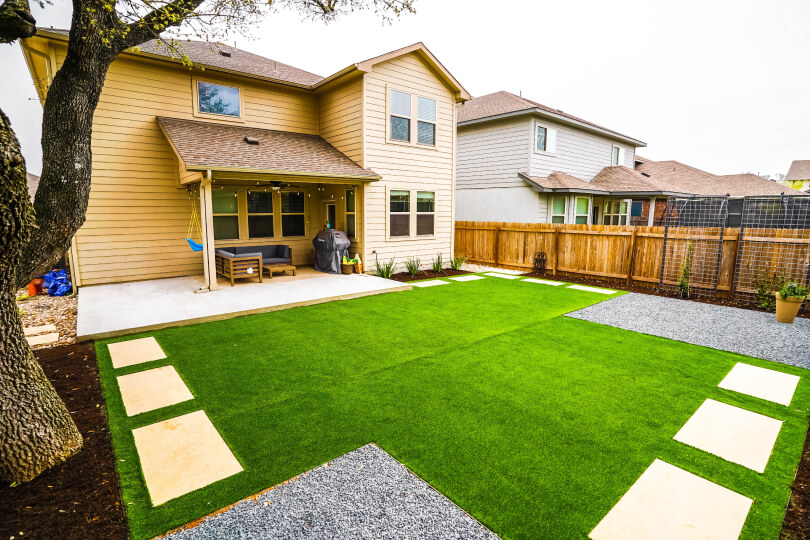 View of a landscaped backyard and the back of a house. There is green grass with white stepping stones. You can see a house in the background.