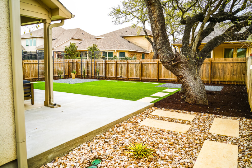 Side view of a landscaped backyard. On the right you can see a patio and on the right there is a large tree. There is green grass in the center of the yard with white stepping stones. The yard is surrounded by a wooden fence.