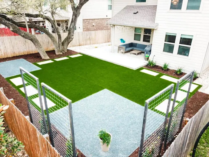 Full view of a landscaped backyard. You can see the back of the house, the patio, a rectangular patch of grass, a square section of pebbles, and white stepping stones around the yard.