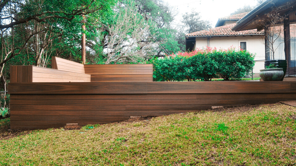 Side view of a wooden deck in a back yard. Grass and shrubbery can be seen in the background.