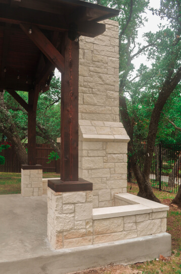 Side view of large white stone fireplace in a backyard. Wooden pillars are connected to the fireplace. Trees in the backyard can be seen.