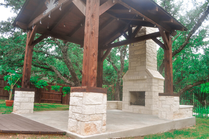Side view of a covered patio in a backyard. The patio features a white stone fireplace. Trees can be seen in the background.