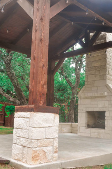 White stone patio with wooden covering. A stone fireplace is built in to the structure. Trees in the surrounding backyard can be seen.