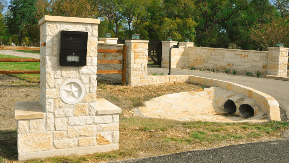 Entrance to a driveway. On the left there is a white stone mailbox and to the right there is a gated entrance surrounded by a white stone wall. There is a grass around the driveway.