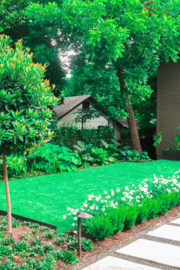 Sideview of a front yard. On the right there is a pathway with white stone steps leading up to the door. The path is lined with white flower plants. You also see a strip of grass and multiple trees in the background.