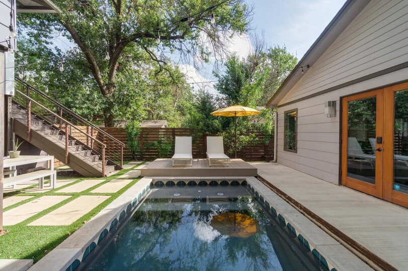 View a pool in a backyard. There is two white pool chairs in the background and you can see wooden stairs on the left side of the pool.