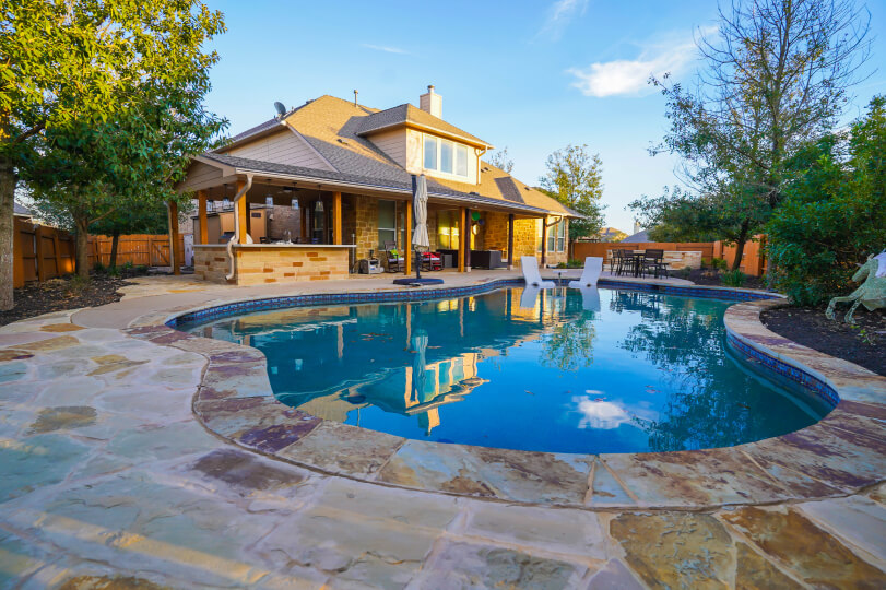 Clear blue in-ground swimming pool with light flagstone around the edges. In the background, there's a house with a large covered patio.