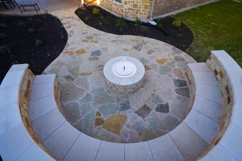 Aerial view of a light flagstone area of a backyard with a stone bench around the perimeter and a fire pit in the middle.