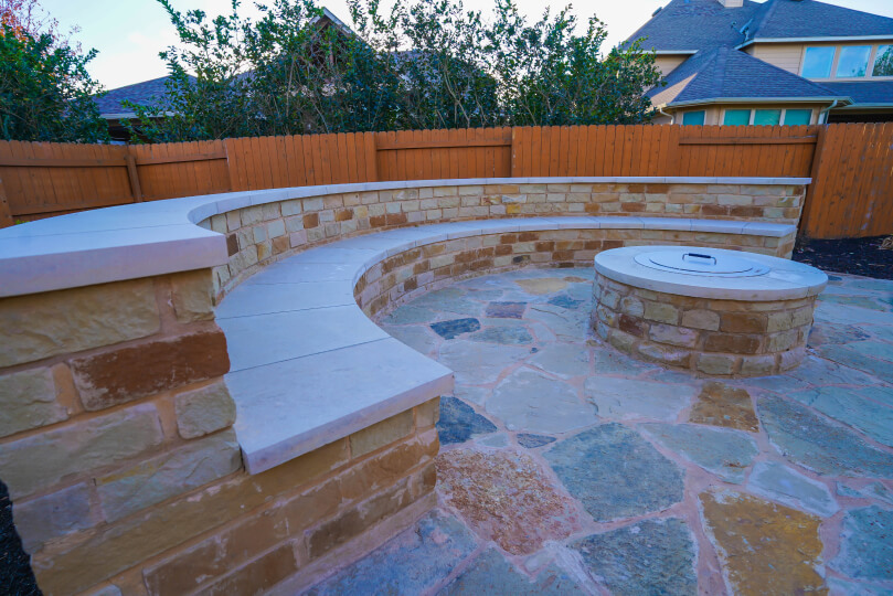 Rounded flagstone area of a backyard with a stone bench around the perimeter and a fire pit in the center.