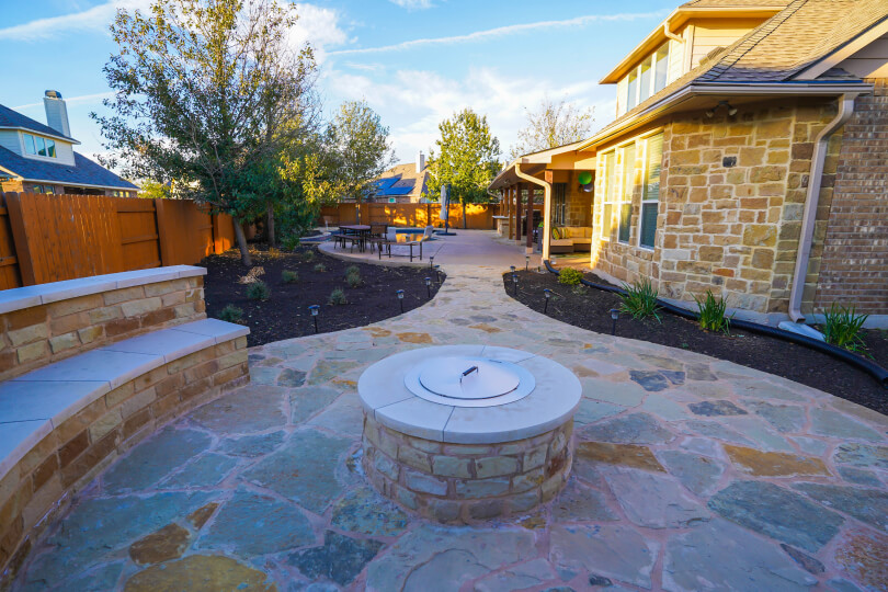 Rounded flagstone area of a backyard with a stone bench around the perimeter and a fire pit in the center.