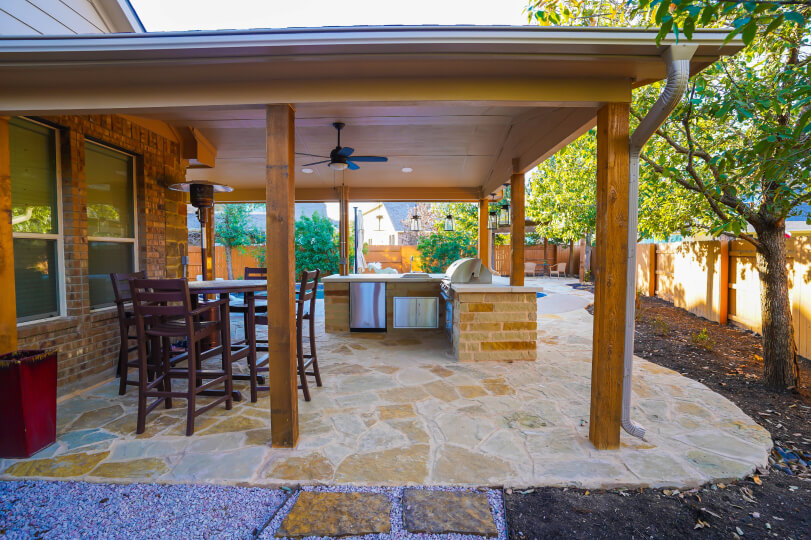 Covered patio with light stone flooring. There's a high top table with 4 barstools around it, and a light stone outdoor kitchen area.