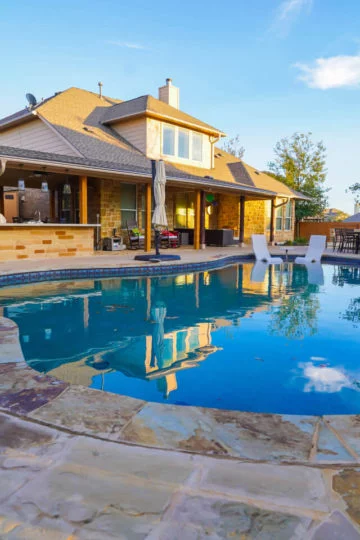Clear blue in-ground swimming pool with light flagstone around the edges. In the background, there's a house with a large covered patio.