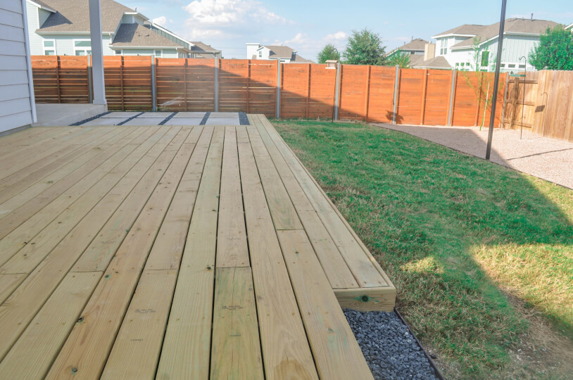 A wooden deck in a grassy backyard. You can see the attached house to the left and there is a wooden fence surrounding the backyard.