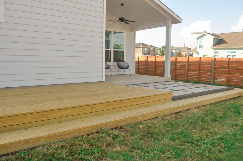 Side view of a back porch. The wooden deck is attached to a white house and you can see green grass and a wooden fence in the background.