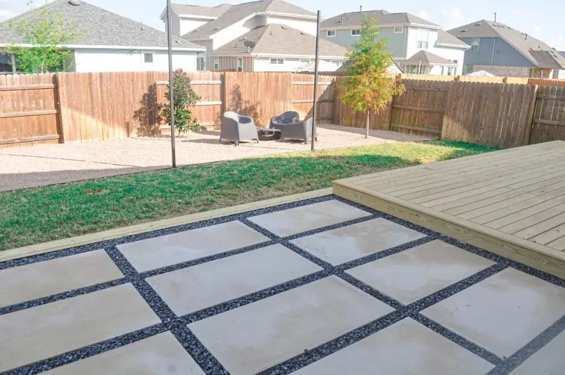 A white stone patio with grey rocks on top of a wooden deck. You can see a grassy backyard with a tree and a wooden fence in the background.