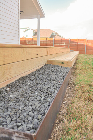 A grey rock bed next to a wooden deck. You can see grass and a wooden fence in the background.