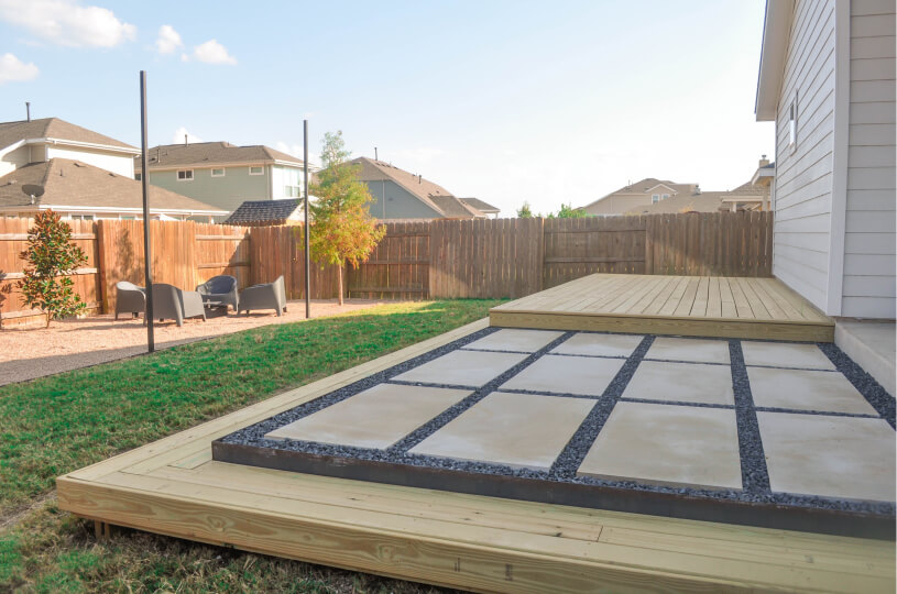 Side view of a stone patio on top of a wooden deck. You can see a grassy backyard surrounded by a wooden fence.