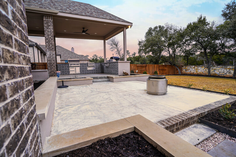 A concrete patio with a small fire pit in the middle. You can also see an outdoor kitchen in the background.