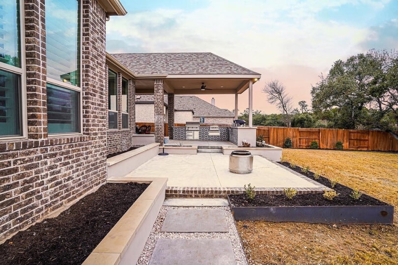 Side view of a concrete patio and built in kitchen in a backyard. You can see plant beds, grass, and a wooden fence in the background.
