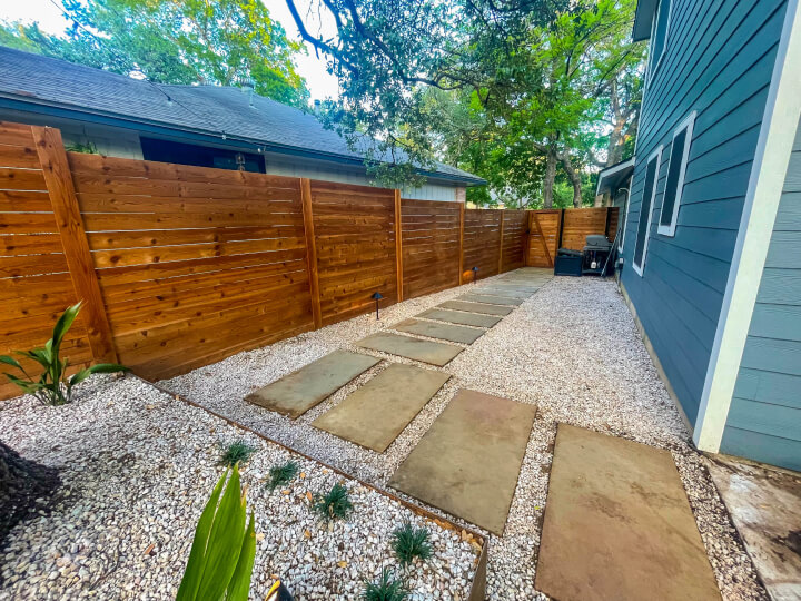 A side yard with a wooden fence around it. There is gravel and stone steps.