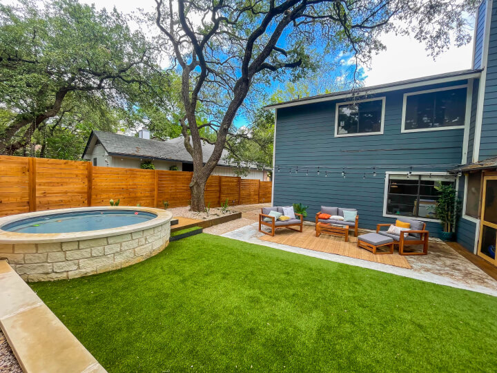 Side view of a landscaped backyard with a view of the back of the house, a small swimming pool and a large tree.