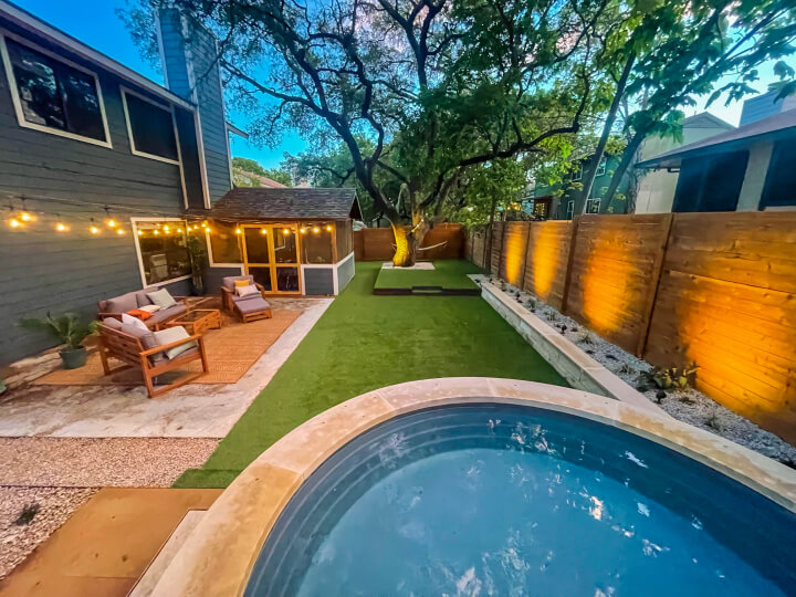 Side view of a backyard with a small pool and green grass. There are strung lights hanging around the yard.