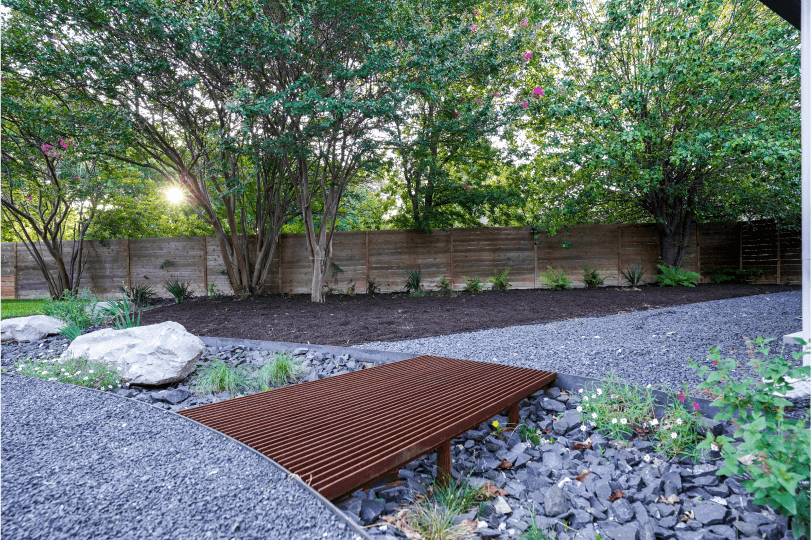 View of a landscaped backyard. You can see a wooden fence and trees in the background.
