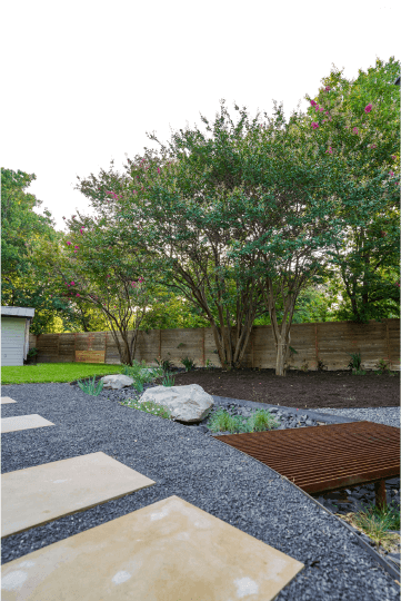 View of a landscaped backyard. You can see a wooden fence and tress and a shed in the background.